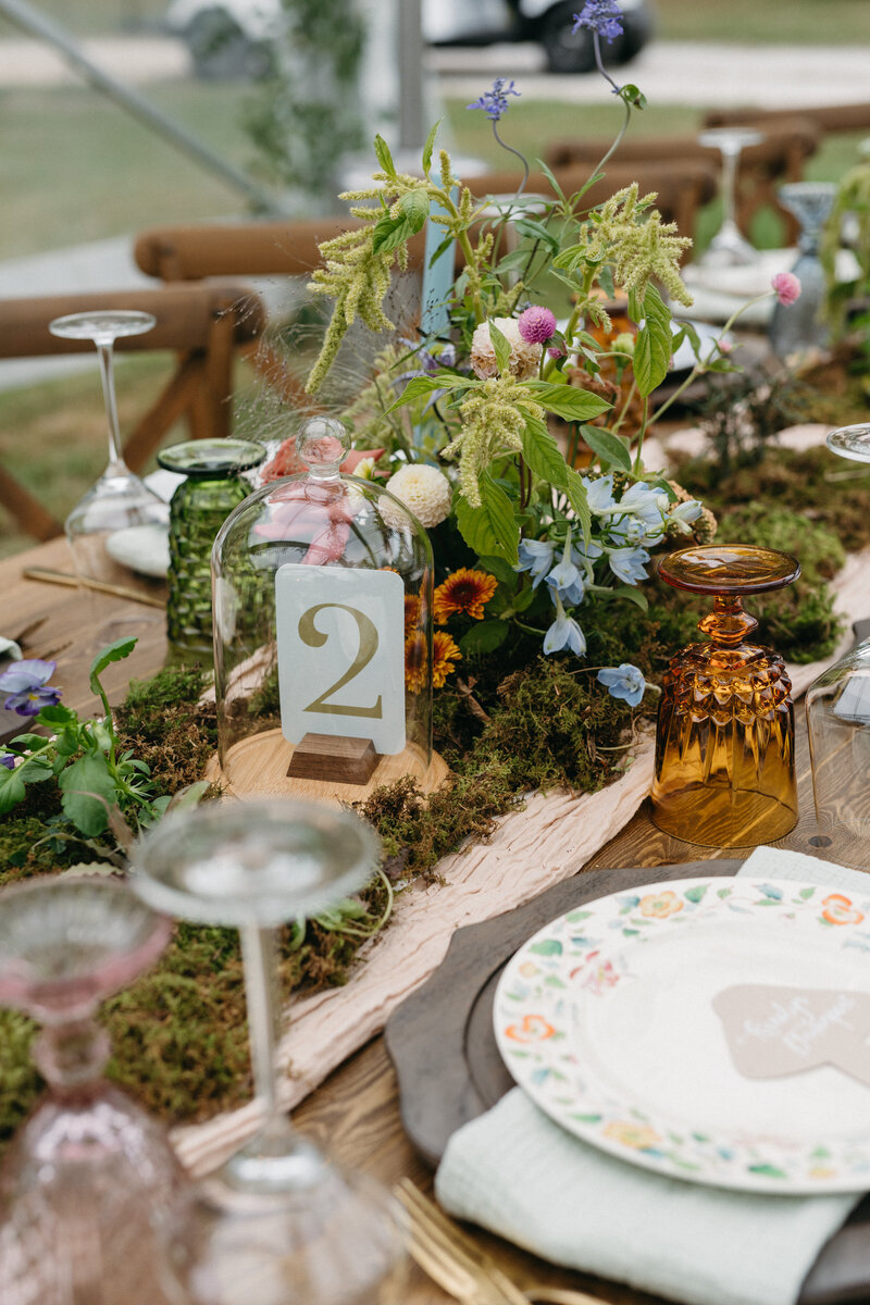Whimsical floral wedding tablescape featuring loose garden blooms in soft pastels, moss-covered runner, vintage glassware, hand-painted plate settings, and table number displayed beneath a cloche dome.
