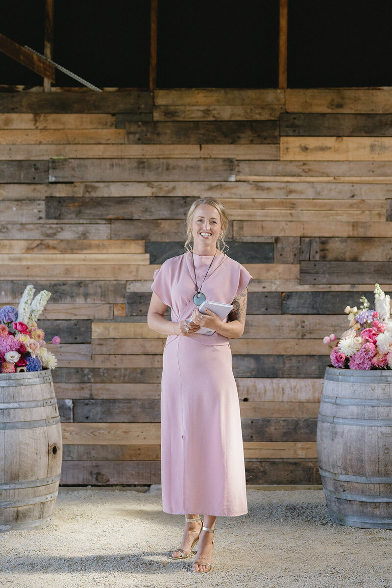 Woman in a blush pink dress and heels standing between two flower-adorned wine barrels, holding a tablet, in front of a rustic wooden wall at a wedding ceremony. By Melissa Clark Photography