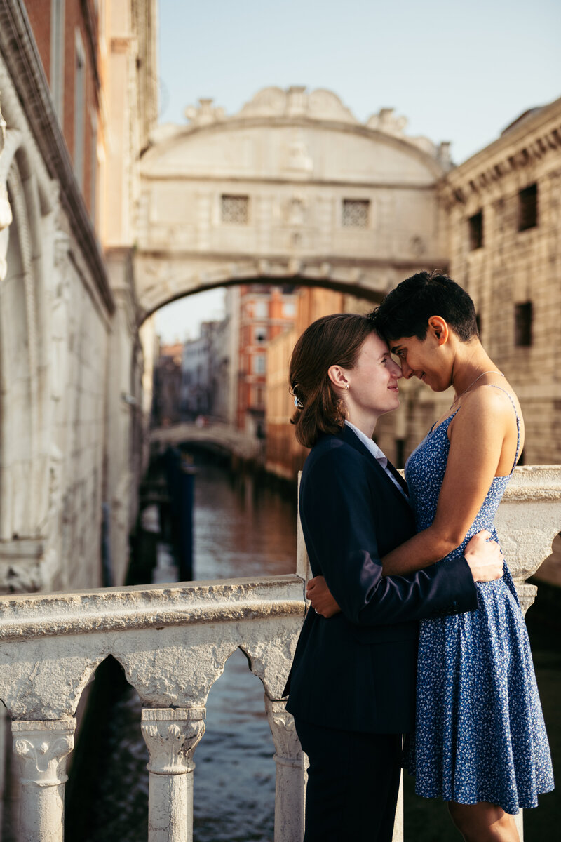 This romantic photo captures the hands of a newly engaged couple in Venice, with the woman's sparkling engagement ring on full display. Against the picturesque backdrop of the city's historic buildings and canals, the image radiates with the joy and love shared between the couple in this perfect moment.