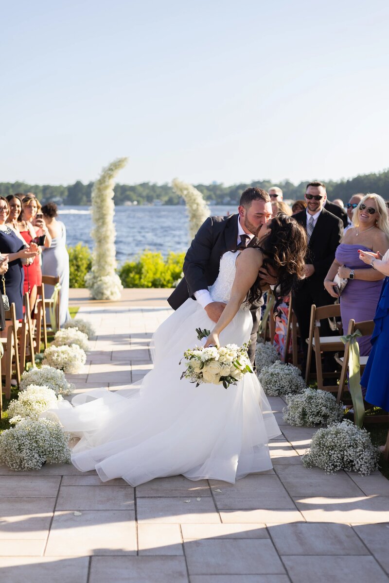 Best Rhode Island Wedding Photographers | Kelsey Sheehan Photography Timeless Rhode Island Weddings | A newlywed couple shares a romantic dip and kiss on a sunlit outdoor aisle, surrounded by joyous guests and white flowers, with a serene lake in the background.