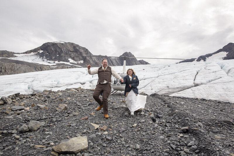 Newlyweds excitingly exiting the helicopter that just landed in front of a glacier with mountains around them.