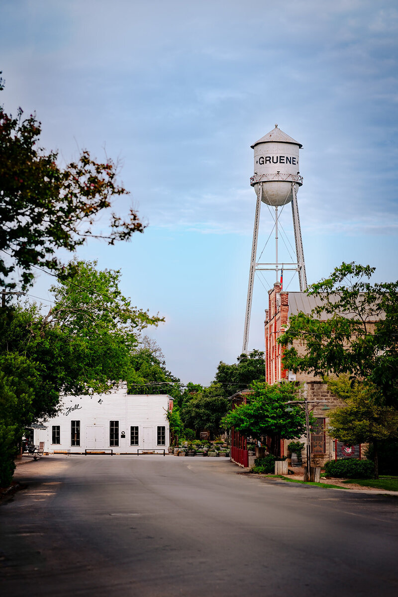 Historic Gruene water tower rising above brick storefronts and green trees in the Gruene Historic District of New Braunfels, Texas, under a soft cloudy sky.