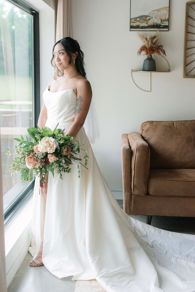 Bride-to-be gazes out of a large panoramic window, with boquet in hand, at the Venue at Mountain Shadow Farm