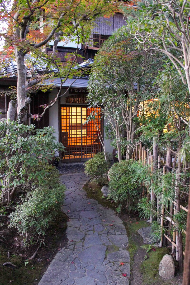 A stone pathway lined with bamboo and fall foliage leading to a traditional home with warm light coming from the doorway in Kyoto, Japan ©Stephanie Dosch | theViatrix Luxury Travel