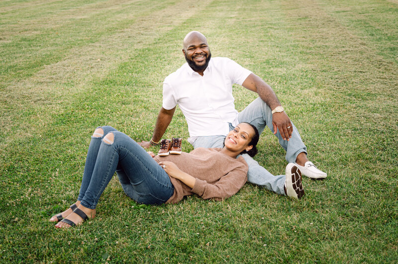 Pregnant couple laying in grass with new baby shoes smiling and laughing