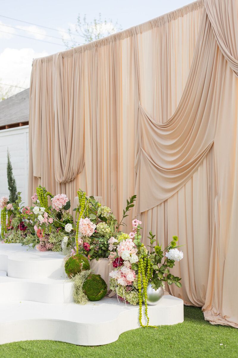 Outdoor stage featuring blush fabric draping, white tiered risers, and spring florals on green turf.
