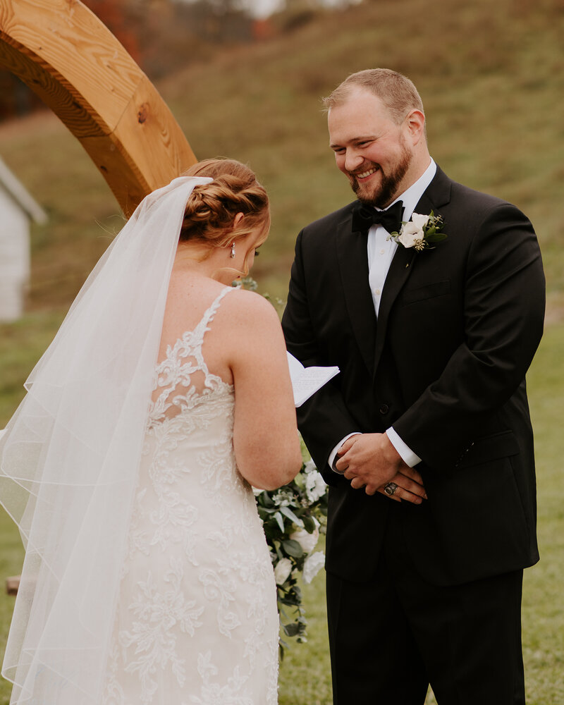 Groom smiling hard as his bride reads her personal vows at the alter during their fall wedding