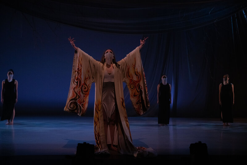 Woman in costume and wearing a mask, facing the audience with her arms stretched towards the ceiling