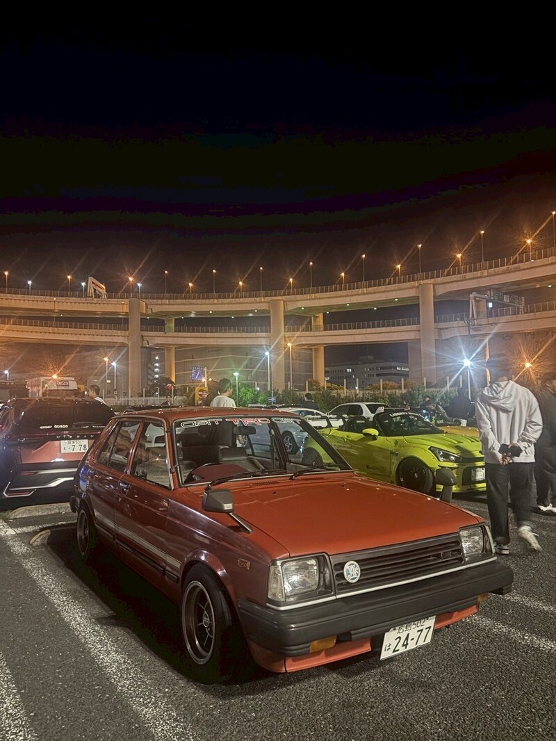 A classic red Toyota Starlet parked among modern sports cars during a vibrant Daikoku Parking Area night meet, capturing the charm of vintage JDM culture.