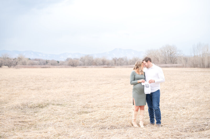 Husband and wife holding a baby outfit together in the open field with mountain views