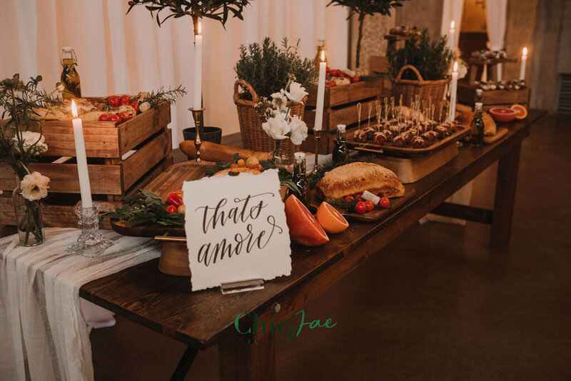 Rustic wooden farmhouse table styled with Italian-inspired charcuterie, candles, herbs, and a hand-lettered “That’s Amore” sign.
