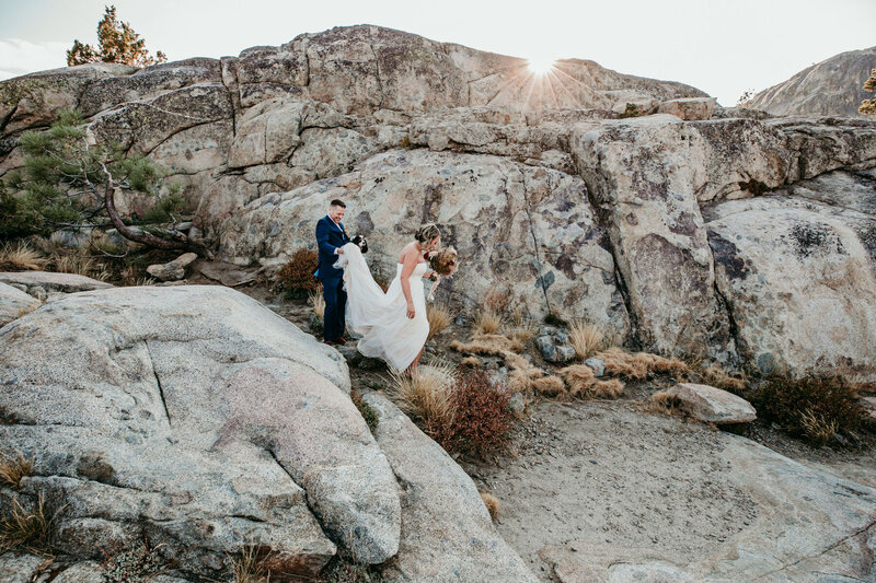 Bride and groom carefully hiking over rocky terrain in Lake Tahoe during their adventurous elopement, with golden sunlight peeking over the mountains in the background.