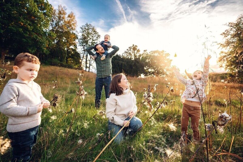 A family of four explores the golden fields of Hudson Valley, NY, with children playing in the wild grass as parents watch over them.