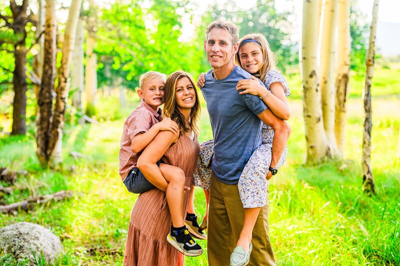 Parents giving kids piggyback ride in summer green aspens and grass smiling and looking at the camera in Flagstaff Arizona
