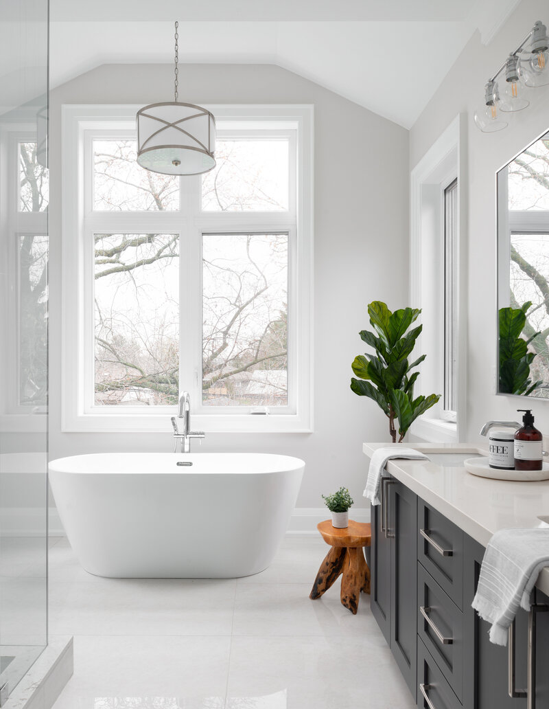 Almost fully white bathroom with a large standalone tub centred under a window, the bathroom has dark blue-grey cabinets, and a large plant in the corner.