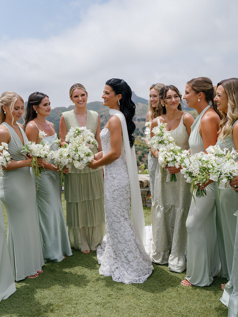 Bride with a sleek, dramatic Kohnur romantic wedding hairstyle (worn down) and luminous soft glam bridal beauty makeup, posing with bridesmaids on a sunny mountain terrace.