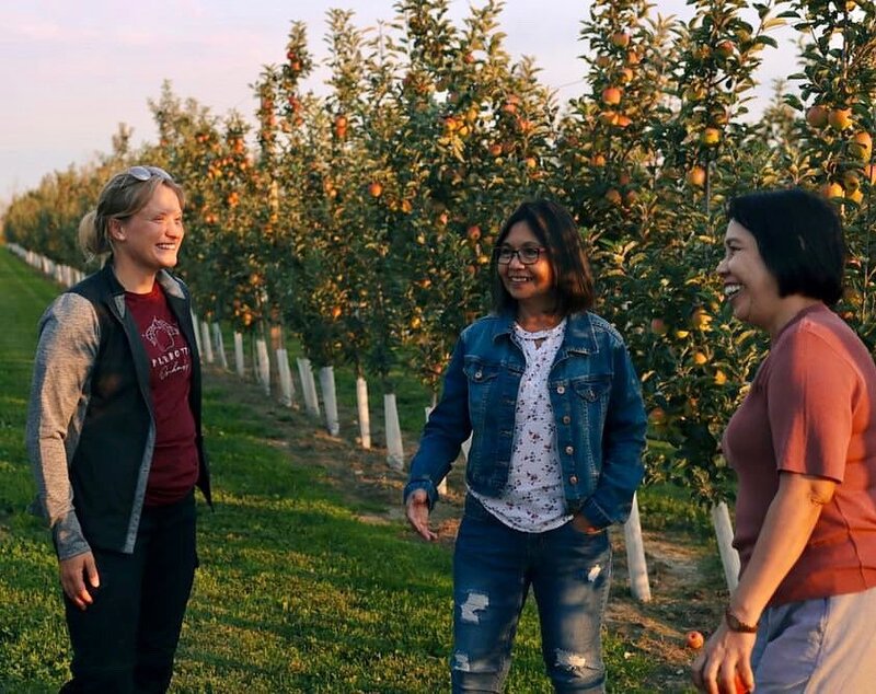 Chelsea, the owner of Applebottom Orchards, chatting with two smiling women in the orchard during golden hour.