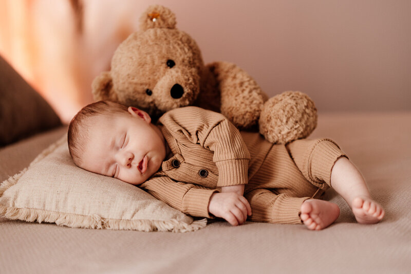 Newborn baby peacefully sleeping in ribbed neutral outfit with a plush teddy bear on a cozy bed — lifestyle in-home photography session in Las Vegas by Angela Wolf Photography