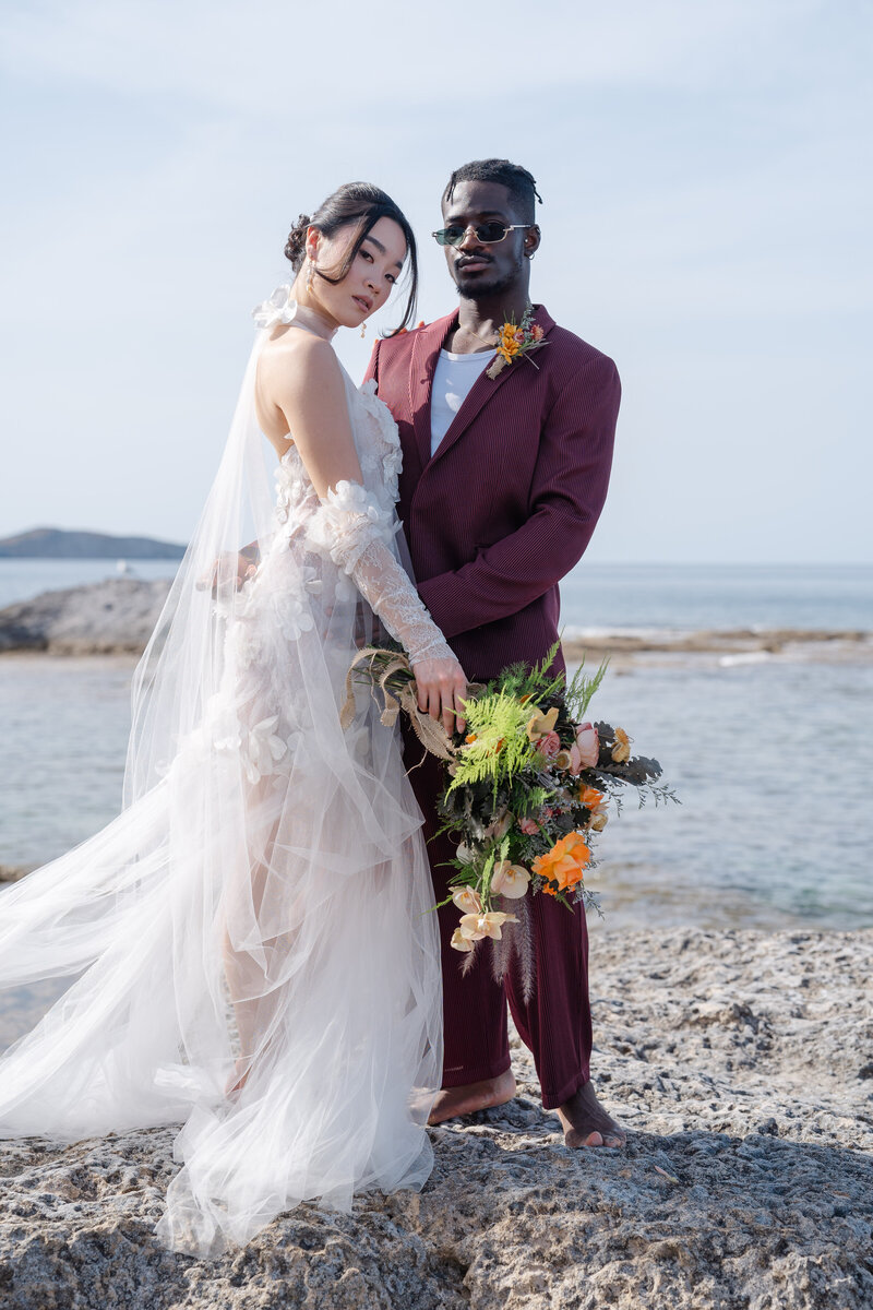 Modern multicultural couple standing barefoot on the coast of Milos, Greece, holding a colourful bouquet – destination wedding by Chi Ramos