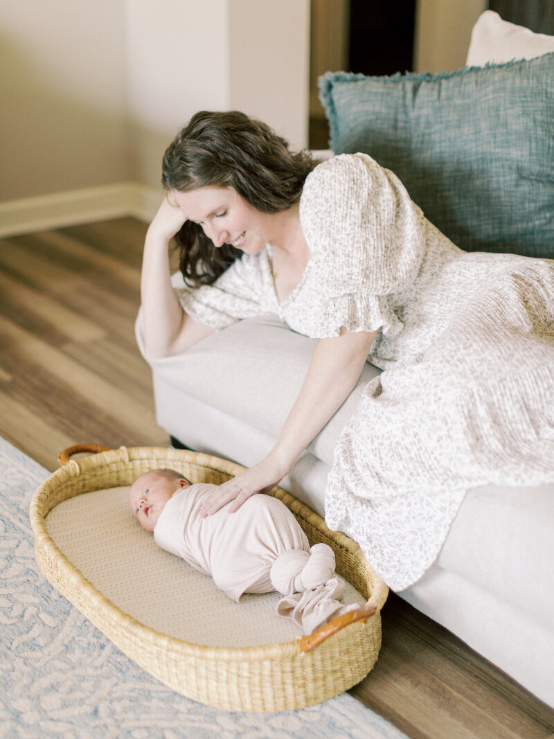 Brunette mother in cream, floral dress lying on couch reaches down to touch her swaddled, newborn son sitting in a moses basket in the floor of their Little Rock home taken by Little Rock newborn photographer Bailey Feeler Photography