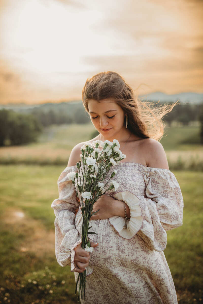 A pregnant mom with her hand on her stomach at sunset, holding flowers, in Tazewell, Virginia.