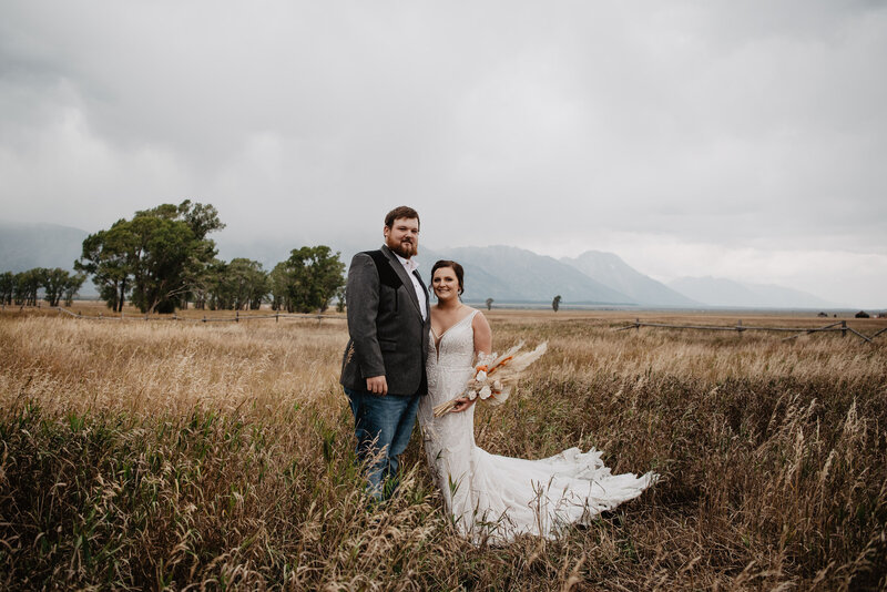 grand teton wedding picture with bride adn groom posing in a fall field with dried grass standing around them while the mountains hide in the fog in the distance captured by jackson hole wedding  photographers