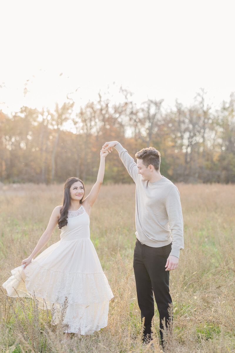 A newly engaged couple dances in a field of tall golden grass in a white dress and sweater