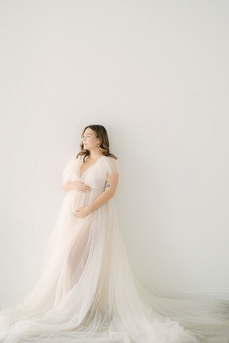 Pregnant mother in a cream tulle dress leans against a wall while holding her baby bump and smiling, Indianapolis portrait Photography