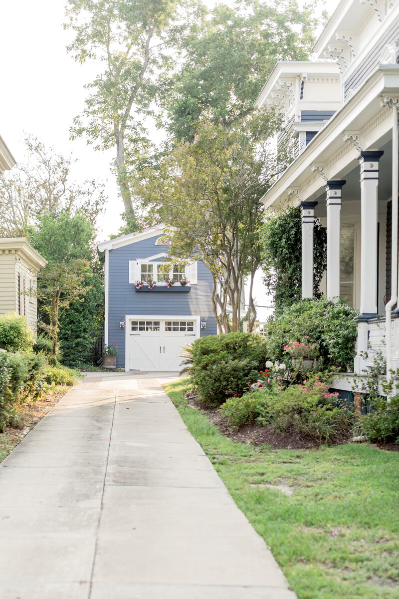 A picturesque blue carriage house with a white garage door, adorned with flower boxes, nestled among lush greenery in a charming neighborhood in downtown Wilmington.