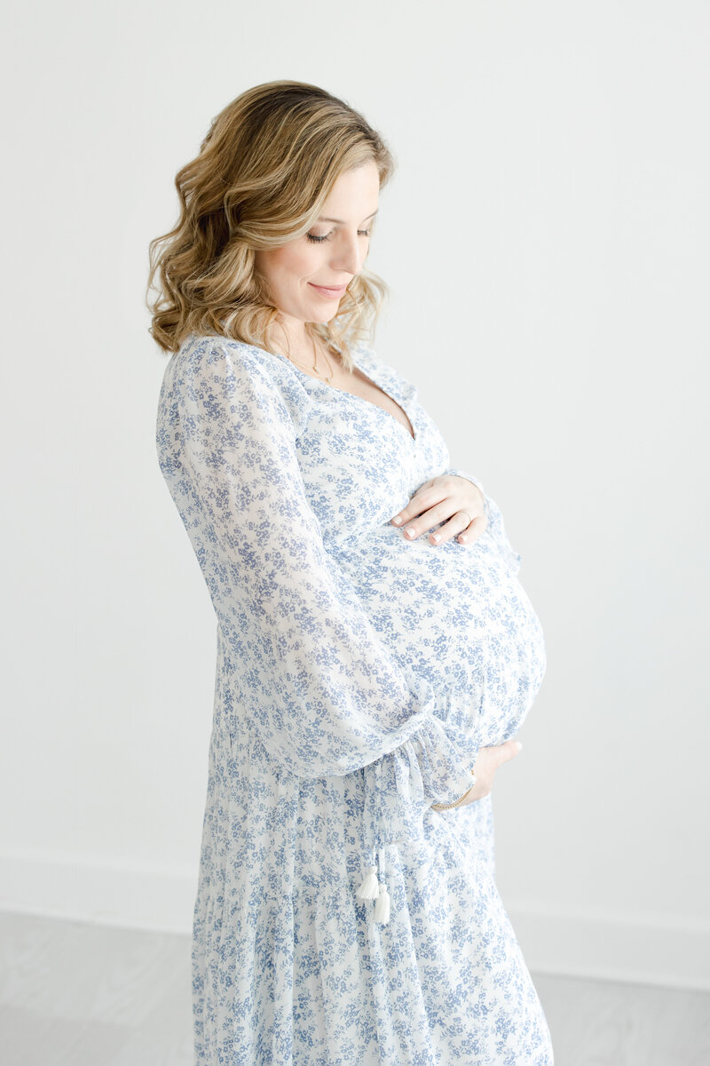Pregnant woman embraces her belly while wearing blue and white floral flowing gown during maternity portrait session