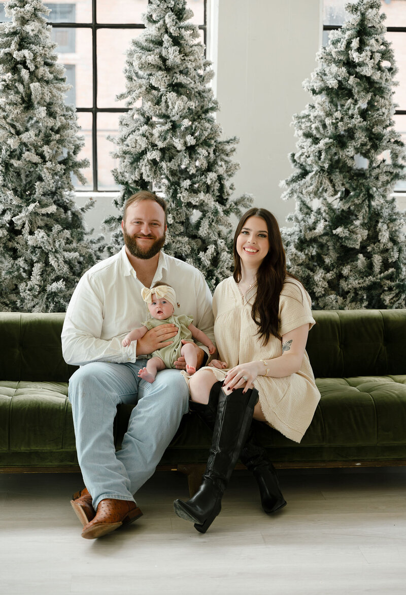 A smiling family sits on a green sofa in front of snowy Christmas trees. The parents hold a baby wearing a headband. The setting is festive and warm.
