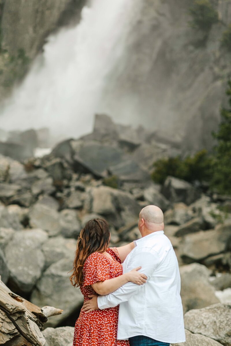 couple embracing near the edge of the cliffside. She is wearing a rust colored lace dress and he is in a beige button up and blue jeans with cowboy boots.