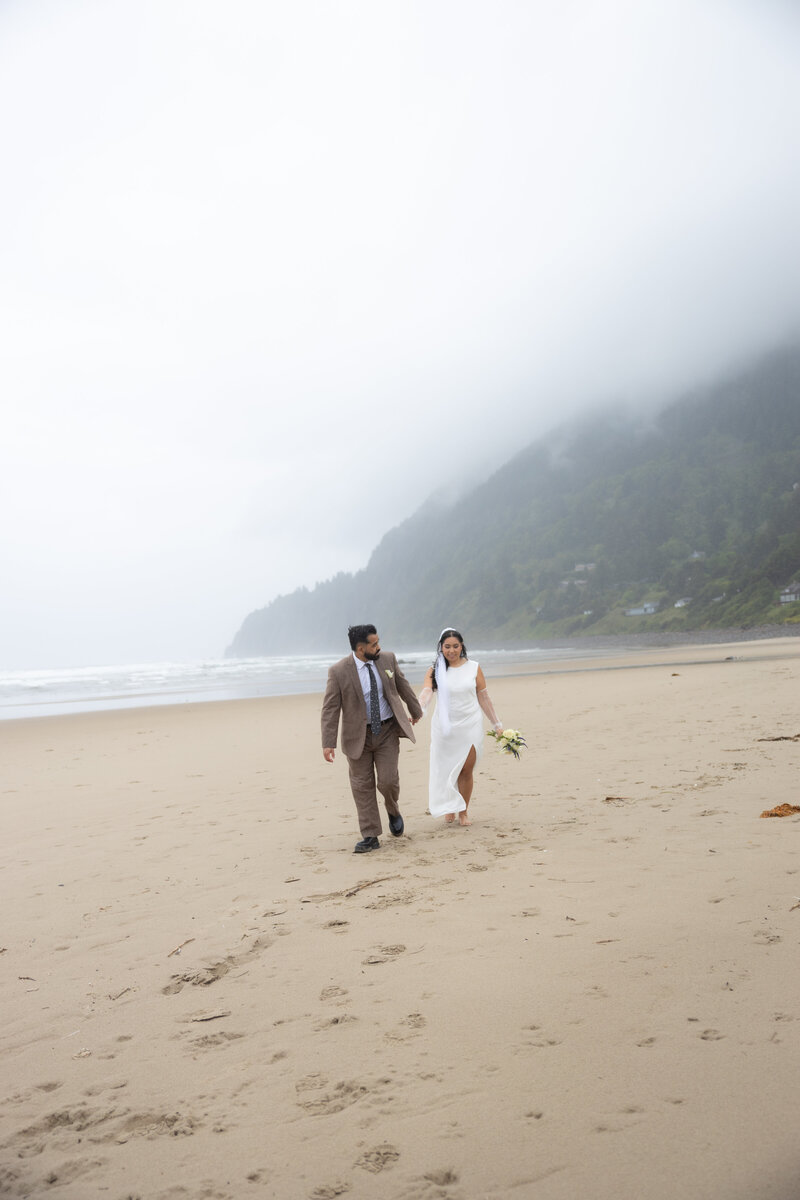 Bride and groom walking barefoot along Manzanita Beach under foggy skies, with mist-covered cliffs in the background during a rainy Oregon Coast elopement.