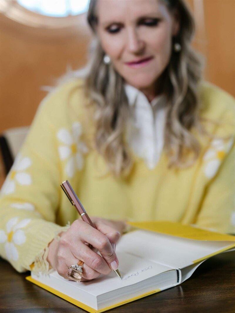Phillipa Leseberg, in a yellow sweater writes in a notebook with a pen, capturing memories for their memoir while seated at a table.