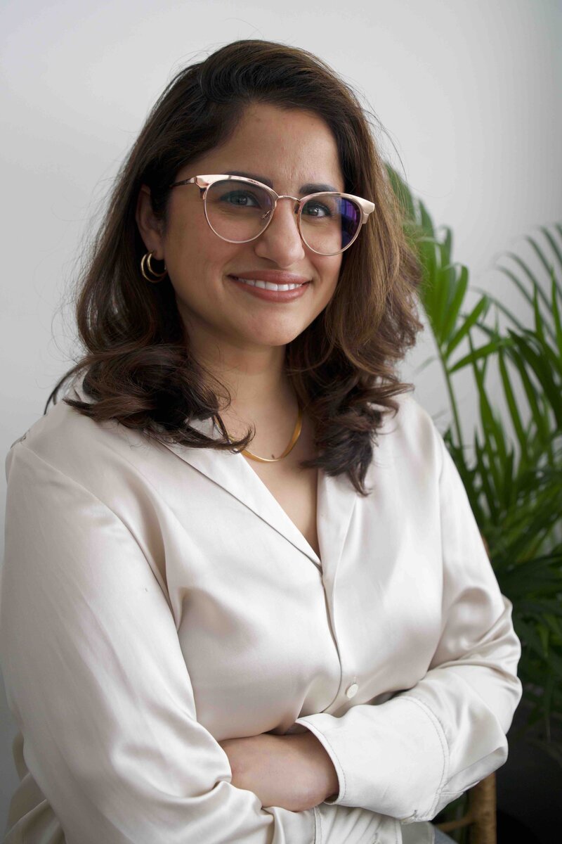 Maya has dark hair, wears a white blouse, and gold rimmed glasses.