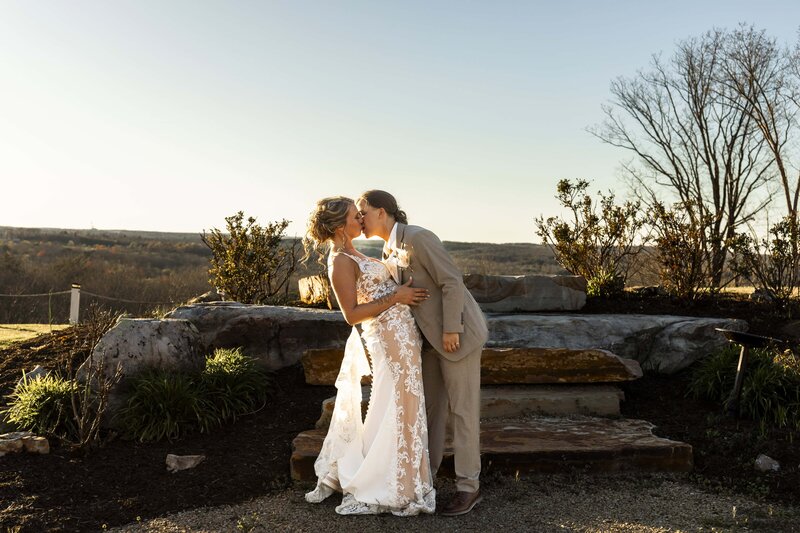 Two brides stand in front of rustic stone steps as the golden hour sun illuminates the landscape around them. One wears a fitted lace gown with a sheer bodice, and the other dons a tailored beige suit. Behind them, the mountaintop views stretch across the horizon, creating a breathtaking setting for their intimate wedding celebration.
