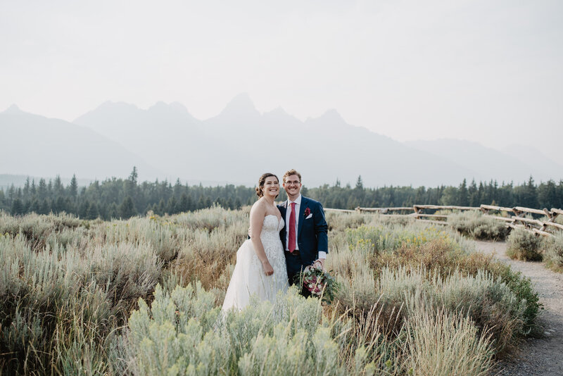 jackson hole wedding  photographer captures bride and groom standing together among sage brush and smiling with the grand tetons in the distance with light fog over them