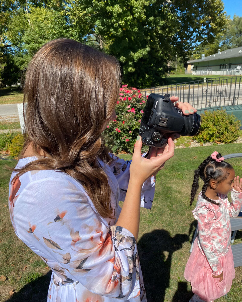 a woman with long brown dark hair standing to the side of the camera with a camera in her hands taking a photo.  trees and young girl in the background