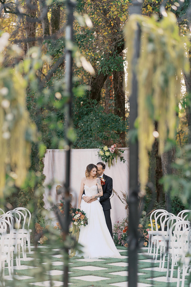 A romantic outdoor wedding ceremony scene at The Greenhouse at Arrows Farm, featuring a bride and groom embracing beneath the trees, surrounded by white chairs, soft florals, and lush greenery—stationery and signage by Grey Cove Designs.