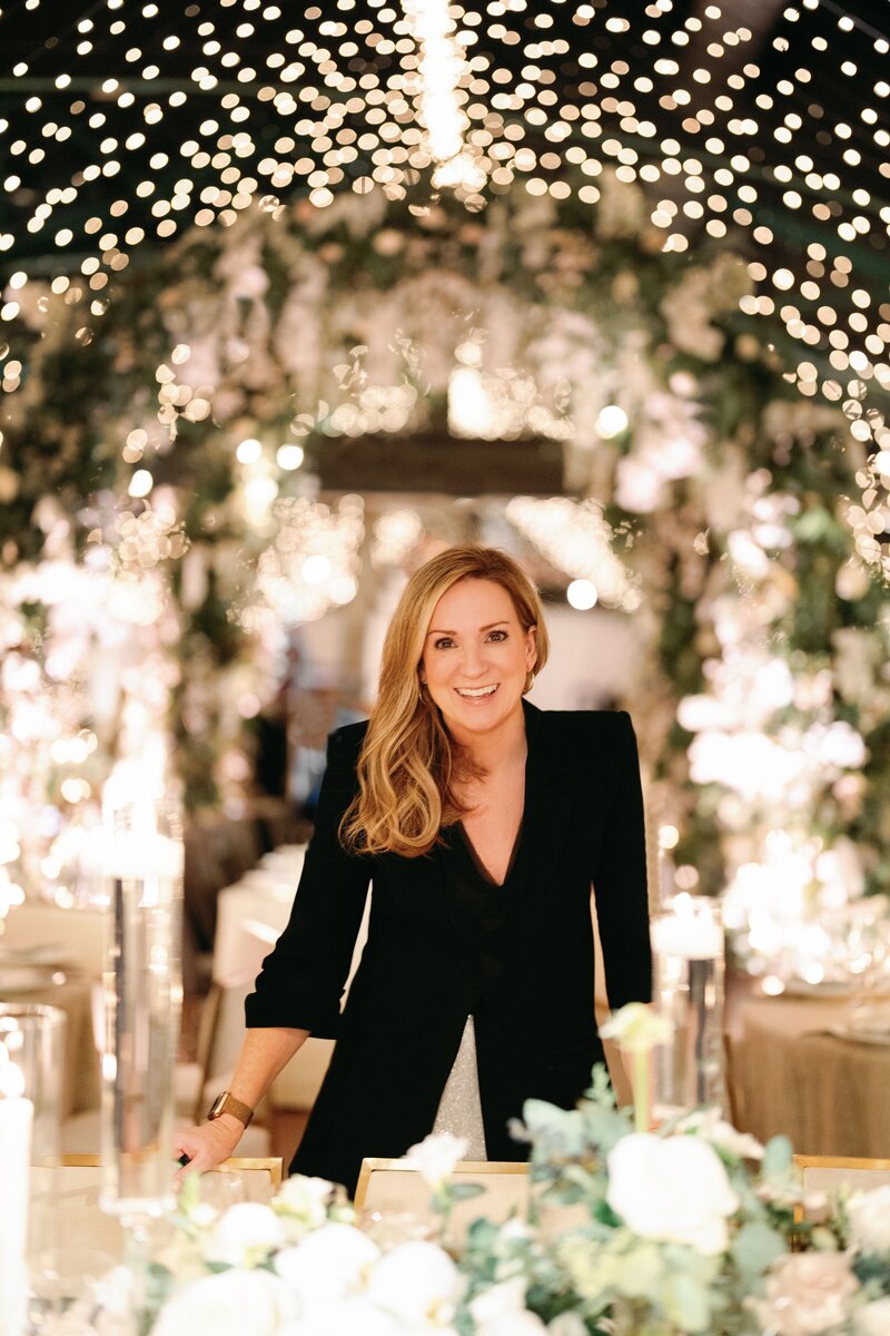 Woman in a black dress smiling at a wedding reception with twinkling lights and floral decorations in the background.