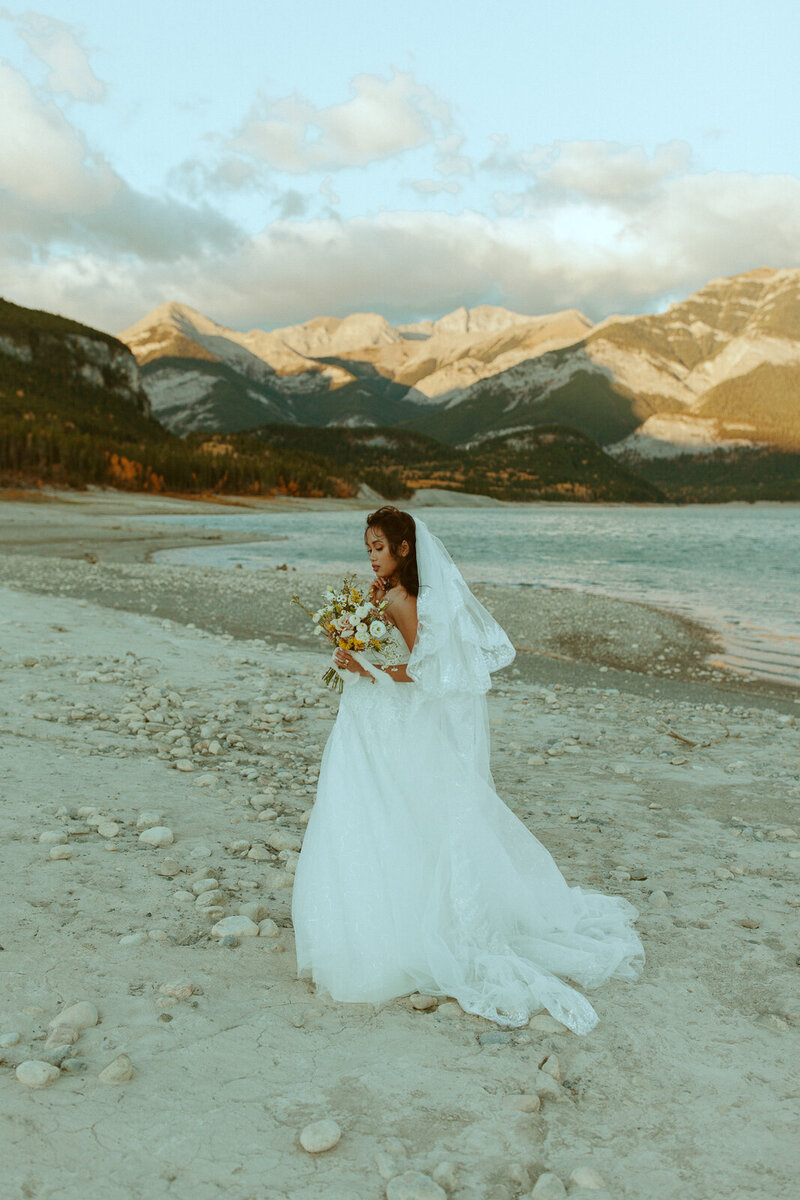 bride looks down at her floral bouquet as wind blows her dress, standing on the shore of barrier lake, in the beautiful kananaskis mountains