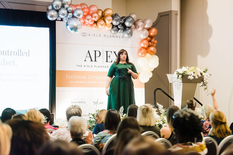 Speaker, Renee Dalo, presents on stage at the Wedding MBA conference in burgundy dress and leopard print belt