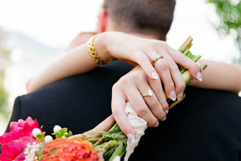 Bride with her arms around the groom's neck holding her bouquet and showing off her ring and jewelry