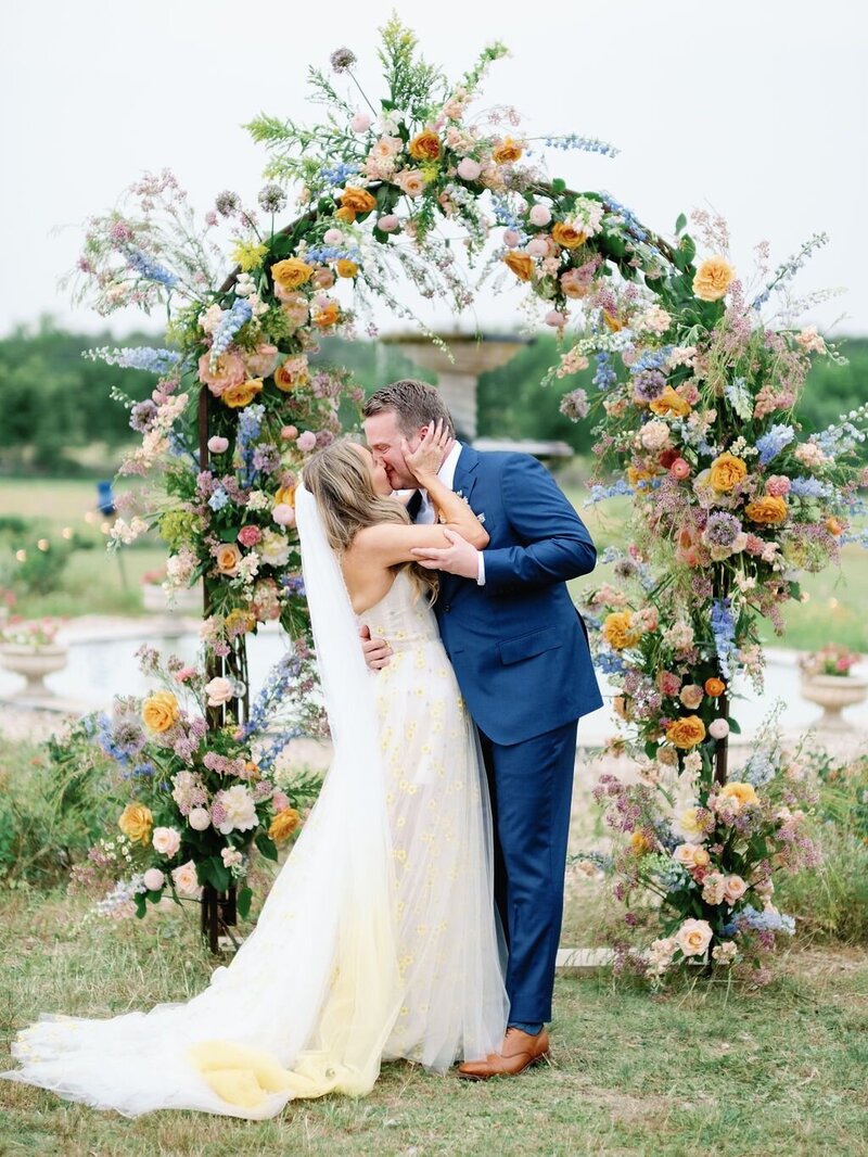 Bride and groom kiss under lush floral arch on their wedding day