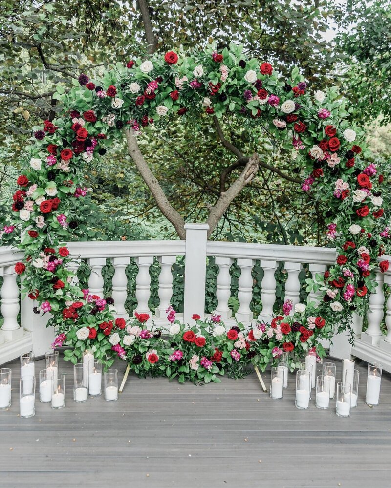 Round floral ceremony arch adorned with red, pink, and blush flowers, set outdoors at the Elkridge Furnace Inn. The arch is framed by lush greenery and a classic white railing, creating a romantic and elegant setting.