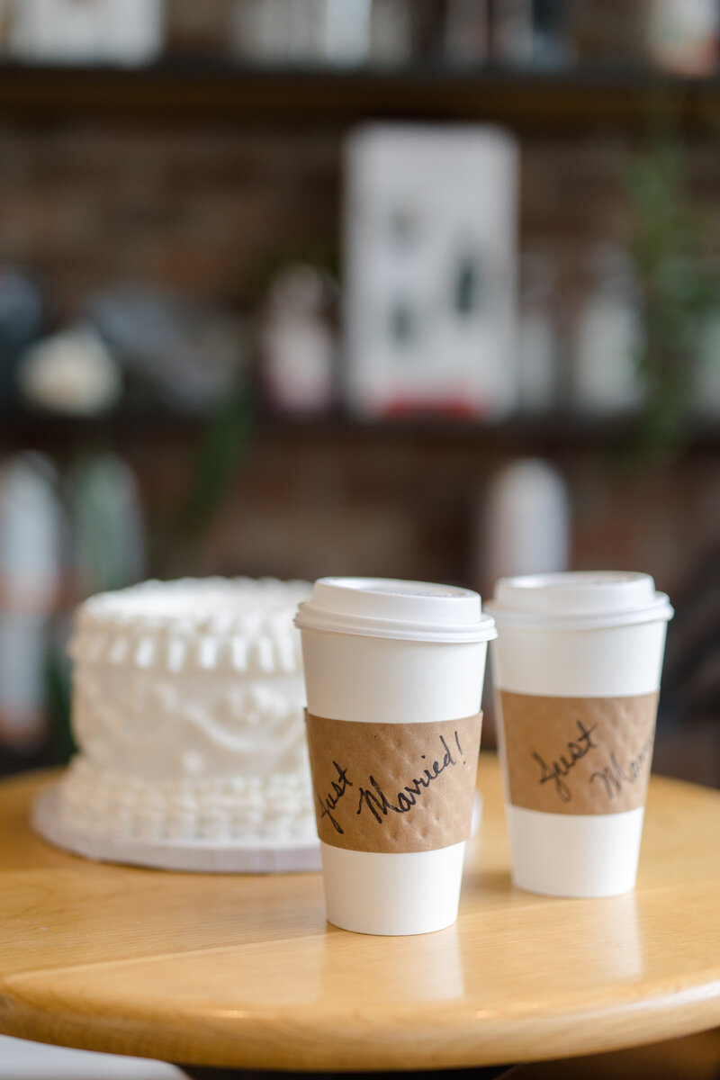 Two paper coffee cups with "Just Married" written on the sleeves on a table in front of a white heart shaped one-tier vintage piped wedding cake at Compass Coffee in Georgetown DC 