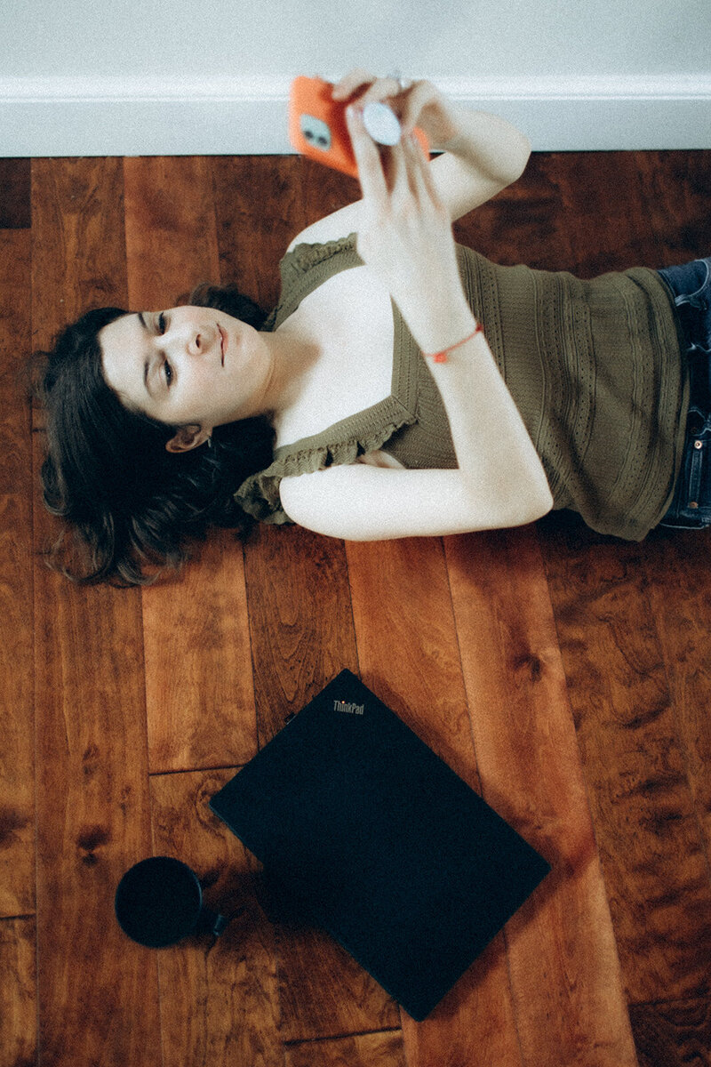 Andrea Shah lying on the floor looking at her phone, with a closed laptop and a coffee mug beside her