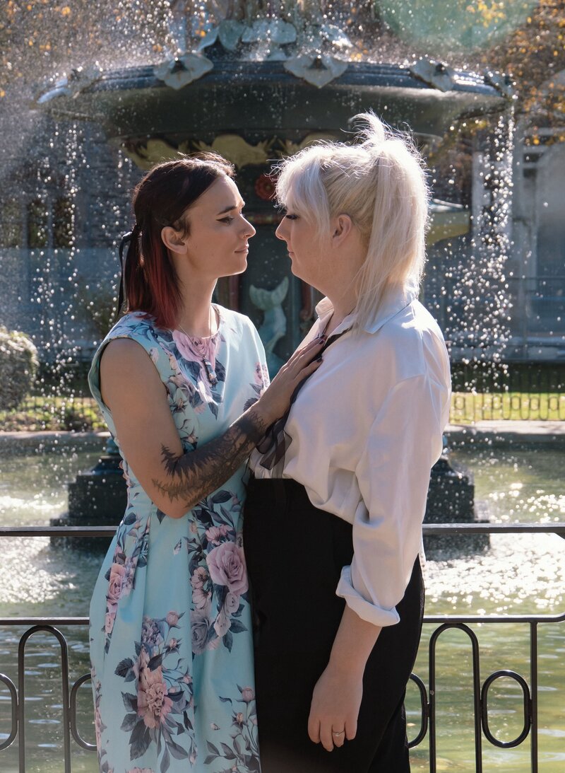 a queer couple celebrates their engagement with photos at Hagley Park in Christchurch, New Zealand. they stand in front of a fountain and look at each other lovingly.