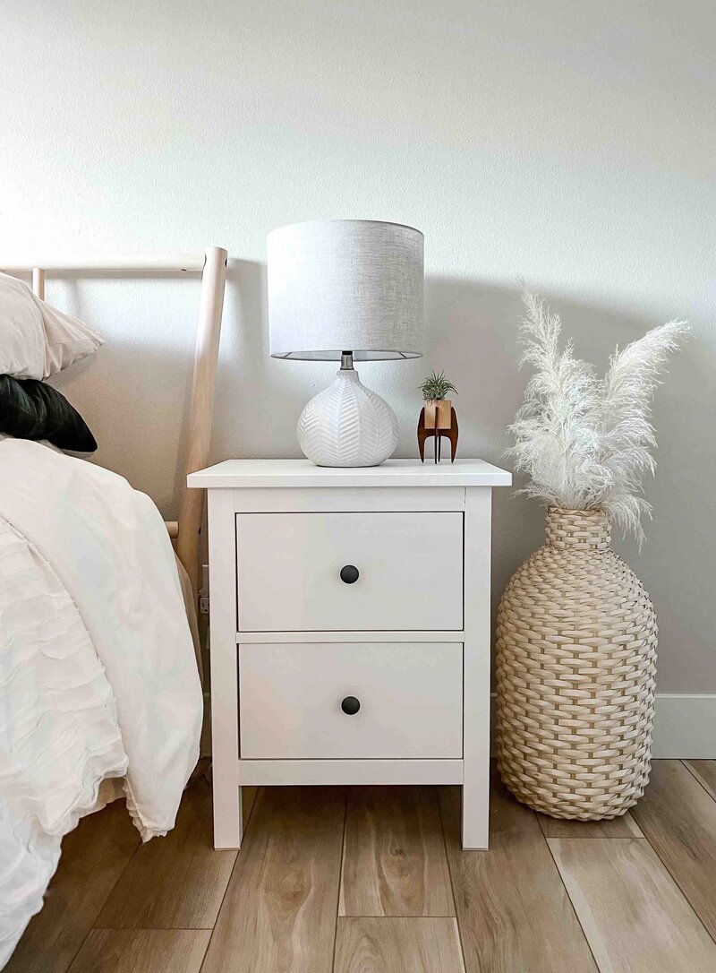 light oak wood floors, beige walls, corner of wooden bed with white bedding, white nightstand with two drawers, white lamp, rattan planter on ground with white pampas grass