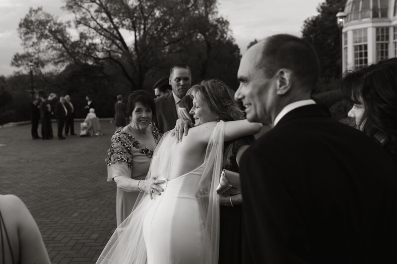 Black and white photo of a bride embracing her mother after the ceremony, surrounded by loved ones, captured by Rainstorm Photo & Video in an emotional, documentary wedding style.